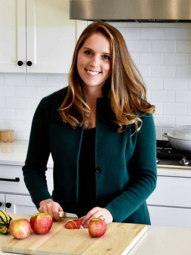 A woman wearing a green blazer chopping an apple on a cutting board.