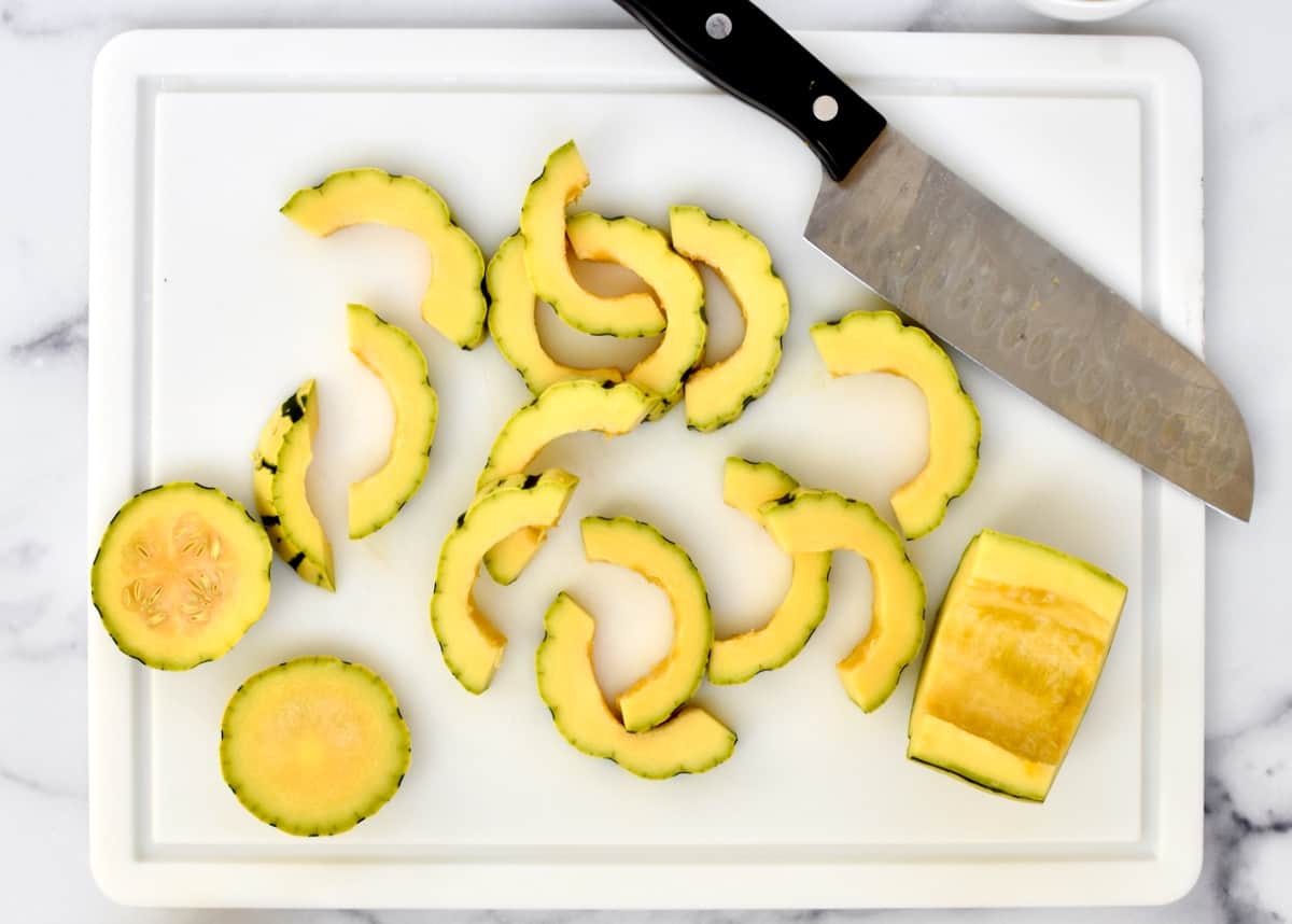 A white cutting board with half moon slices of delicata squash and a knife. 