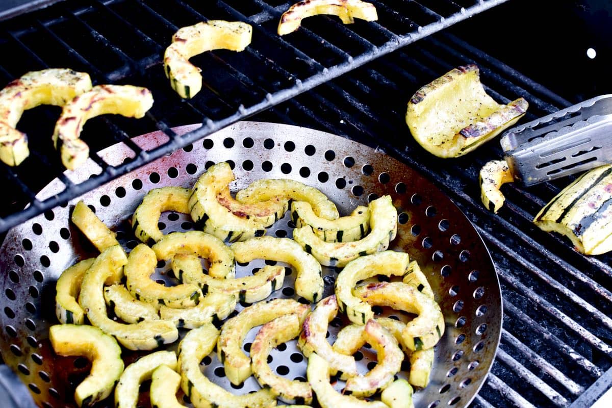 A grill showing delicata squash in a grill wok, and slices directly on the grill grate being turned with a metal tongs. 