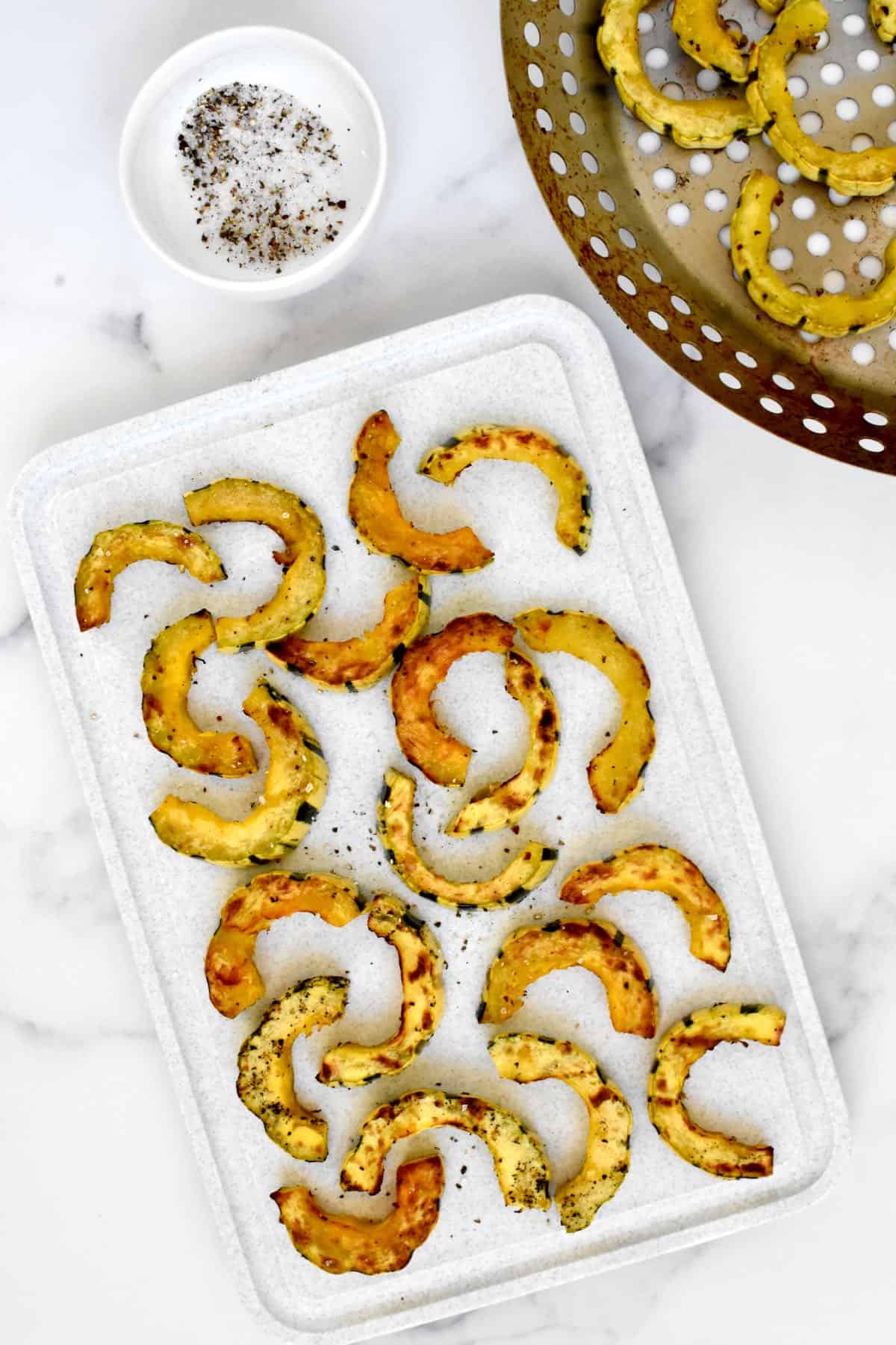 Grilled golden brown squash half moons on a white background, sprinkled with salt and pepper, with a dish of salt and pepper and a grill wok with more squash to the side. 