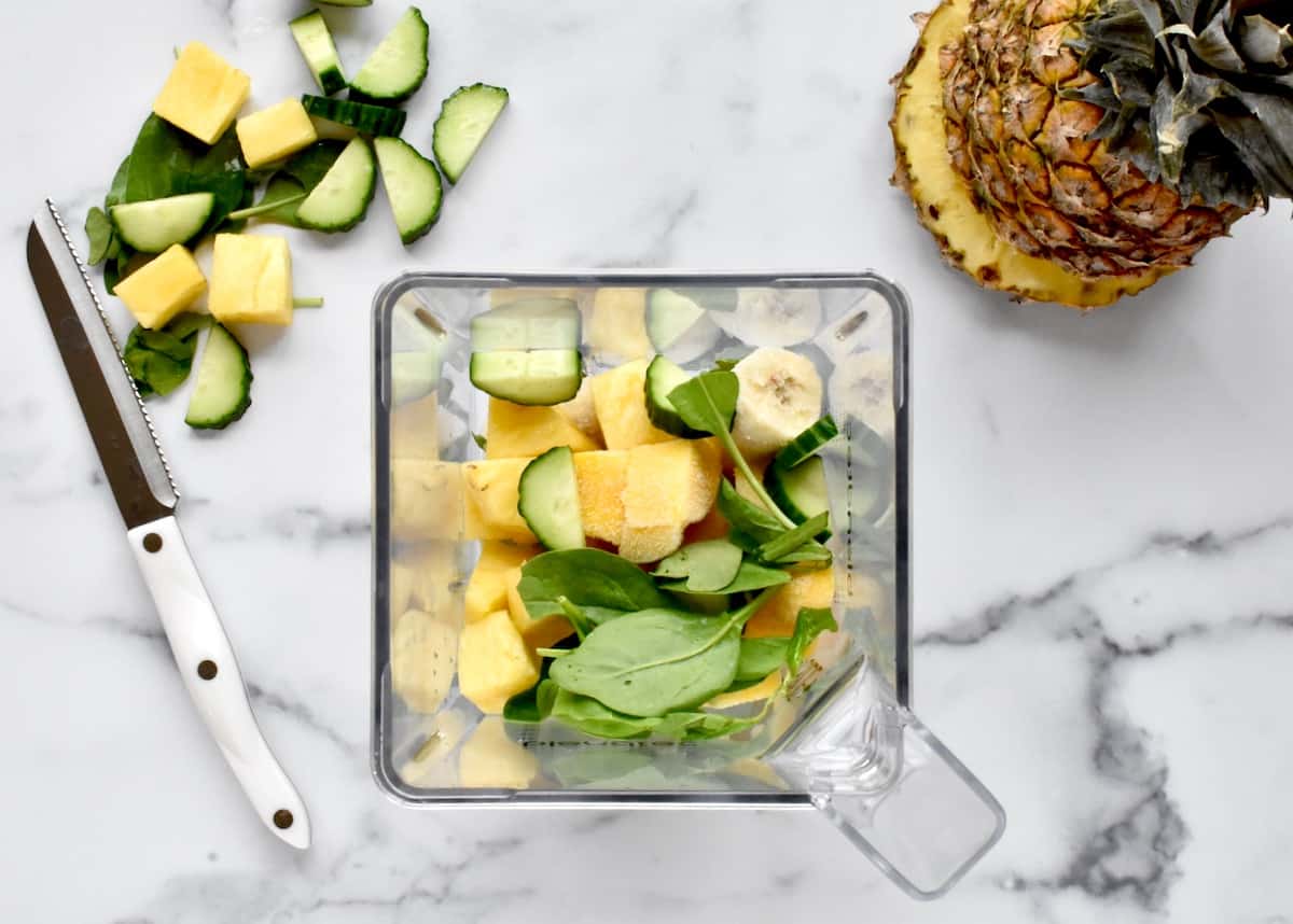 A blender holding unblended pineapple, mango, spinach and cucumber, with a pineapple and chopped fruits and veggies to the side. 