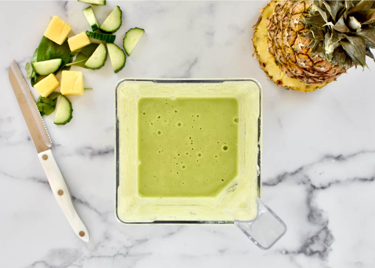 A blender holding blended green smoothie, with a pineapple and chopped fruits and veggies to the side. 