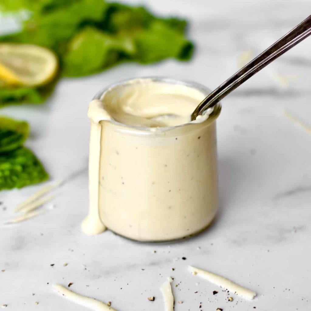 A glass dressing cup with creamy Caesar dressing, with a spoon sticking out and dressing dripping down the side. Romaine lettuce, lemons and parmesan cheese are at the sides.