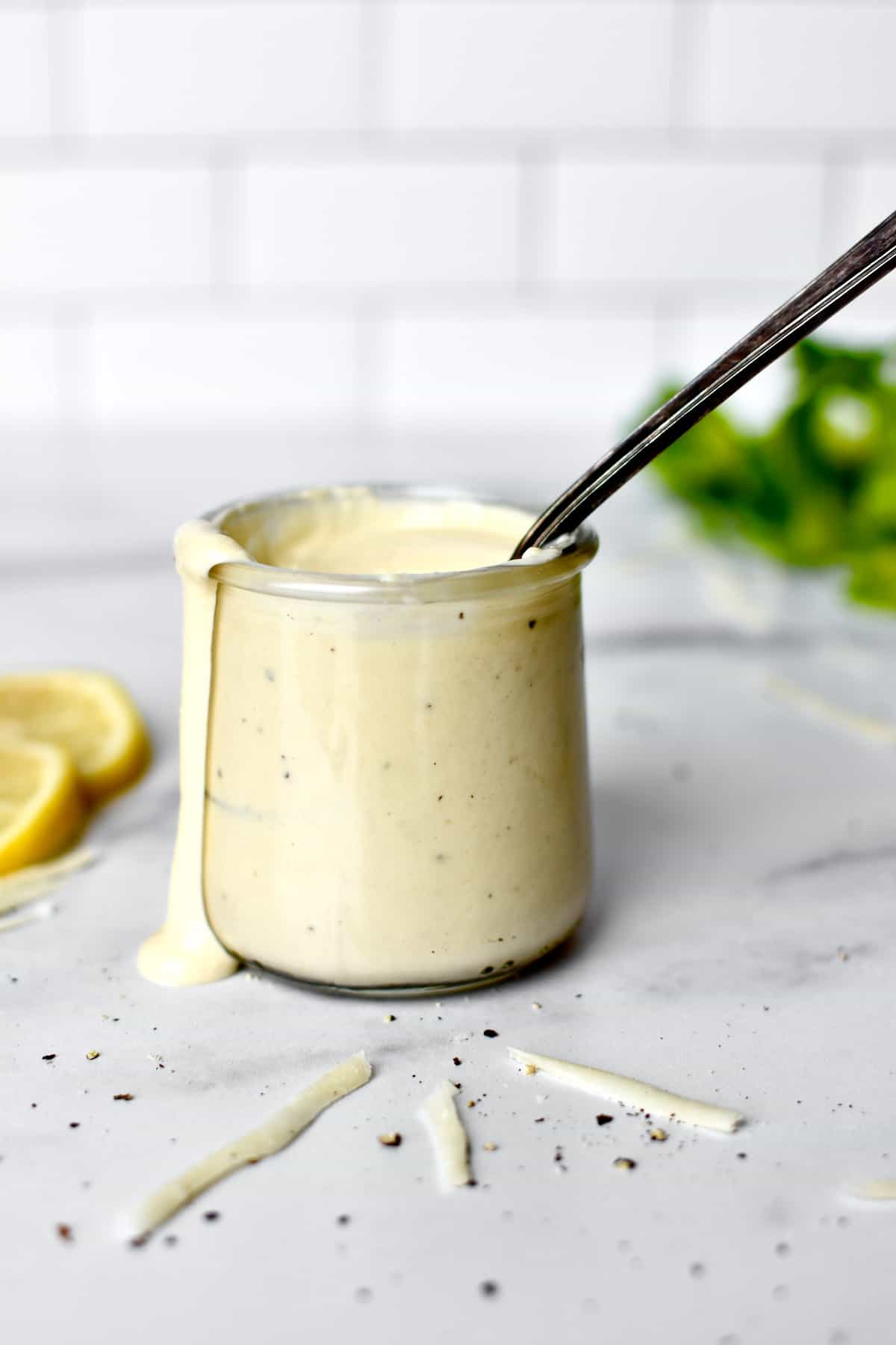 A glass dressing cup with creamy Caesar dressing, with a spoon sticking out and dressing dripping down the side. Romaine lettuce, lemons and parmesan cheese are at the sides.