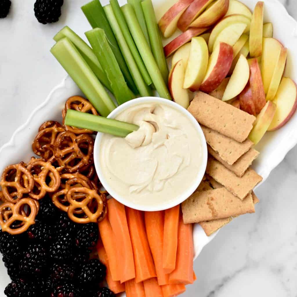 A platter with veggies, fruits, pretzels, graham crackers and peanut butter greek yogurt dip.