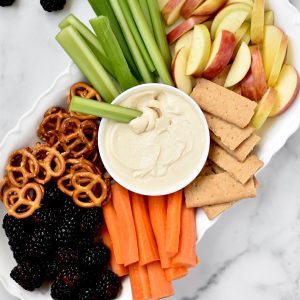 A platter with veggies, fruits, pretzels, graham crackers and peanut butter greek yogurt dip.