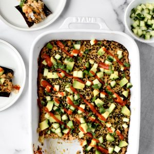 A white casserole dish with sushi bake drizzled with sriracha and topped with avocado and cucumber. A dish of cucumbers and two plates of sushi bake are to the side.