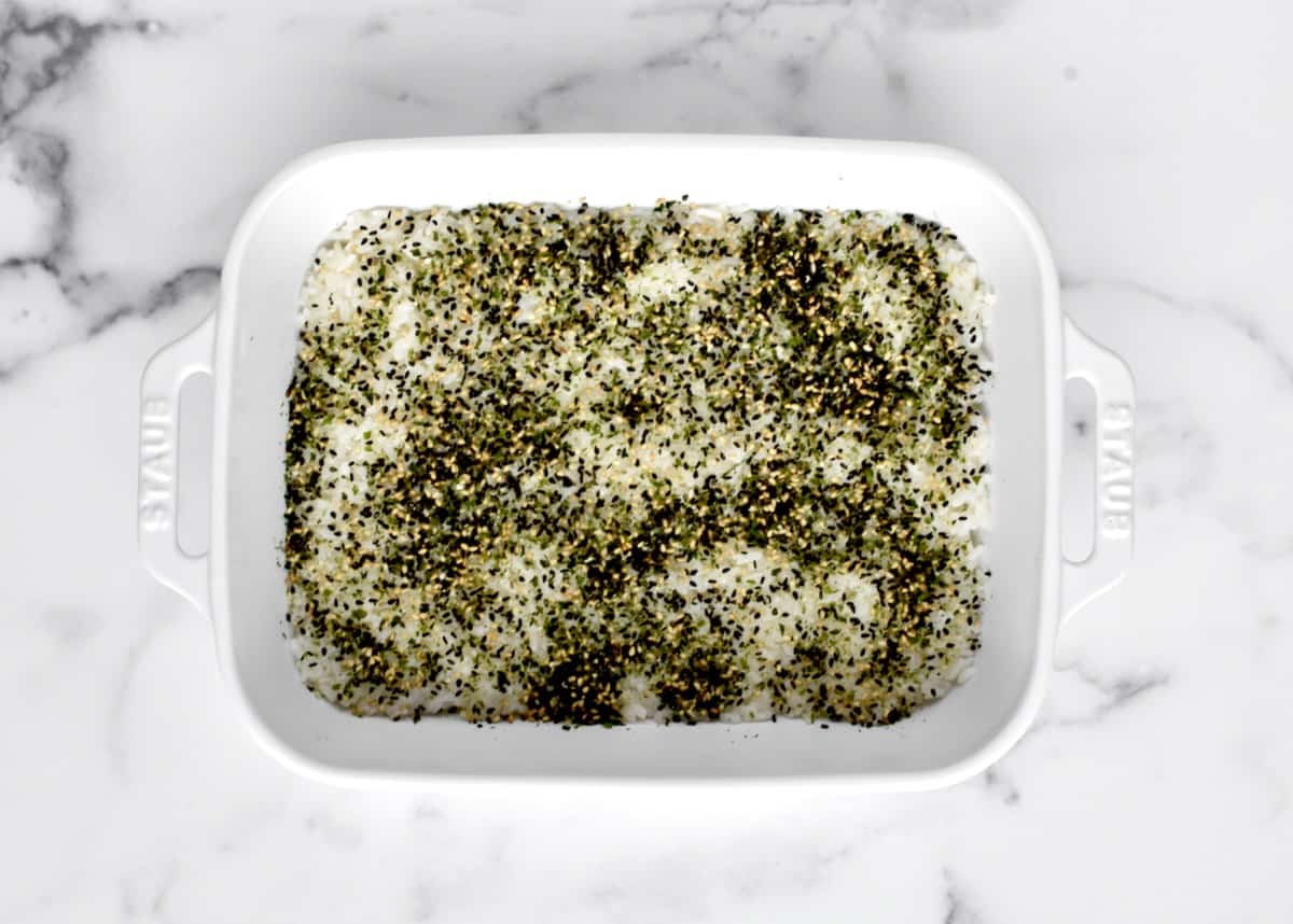 A white casserole dish with a layer of white sushi rice covered in furikake.