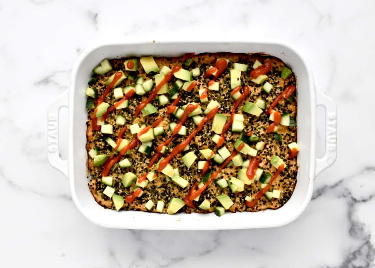 A white casserole dish with baked spicy salmon sushi topped with avocado, cucumber and a sriracha drizzle.