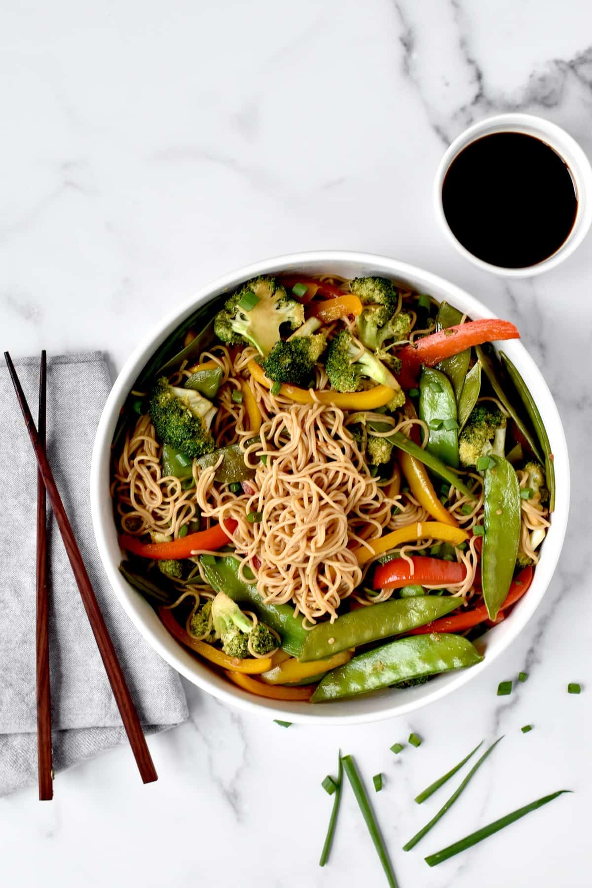 A bowl of ramen noodles with vegetables in stir fry sauce, with a side of soy sauce and two chopsticks to the side.