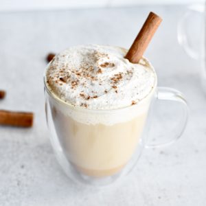 A glass coffee mug with a latte, topped with whipped cream and pumpkin spice. A cinnamon stick garnishes the latte.