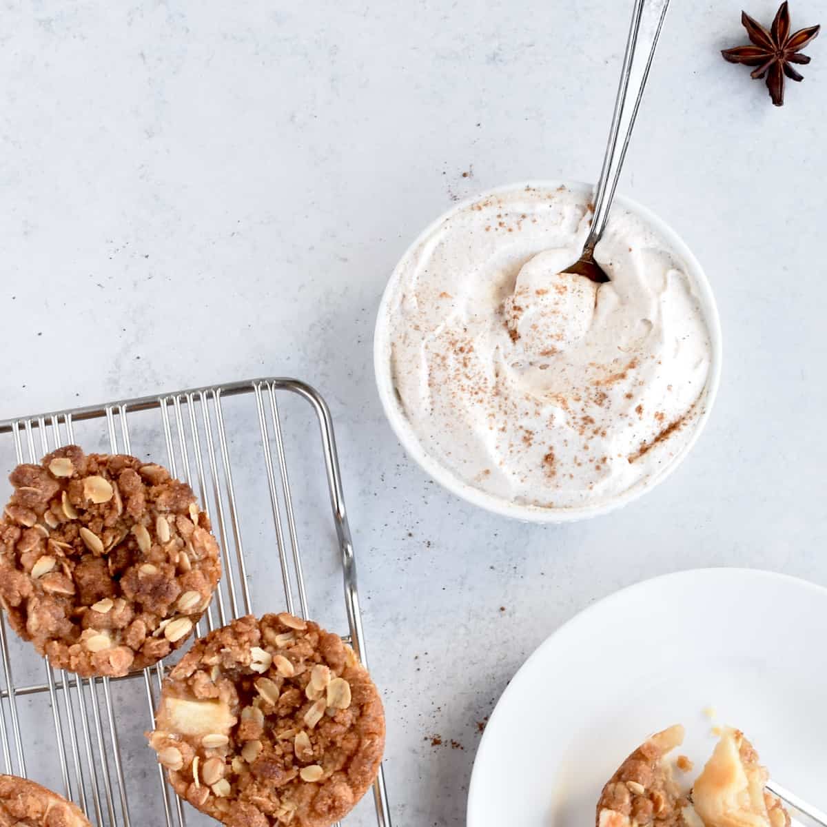 A white bowl of pumpkin spice whipped cream on a grey background. Mini apple crumbles are on a cooling rack and white plate to the side.