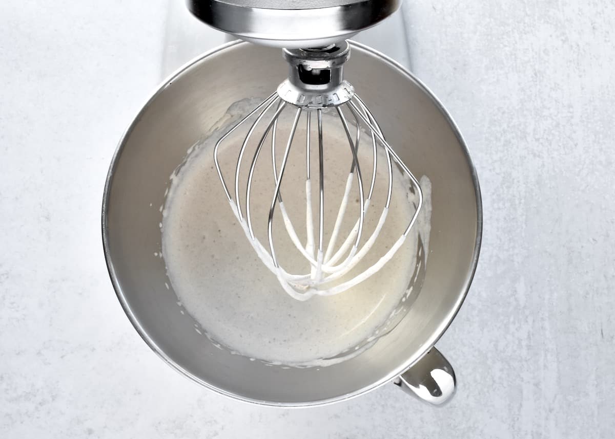 The stainless steel bowl of a stand mixer with cream at no peaks.