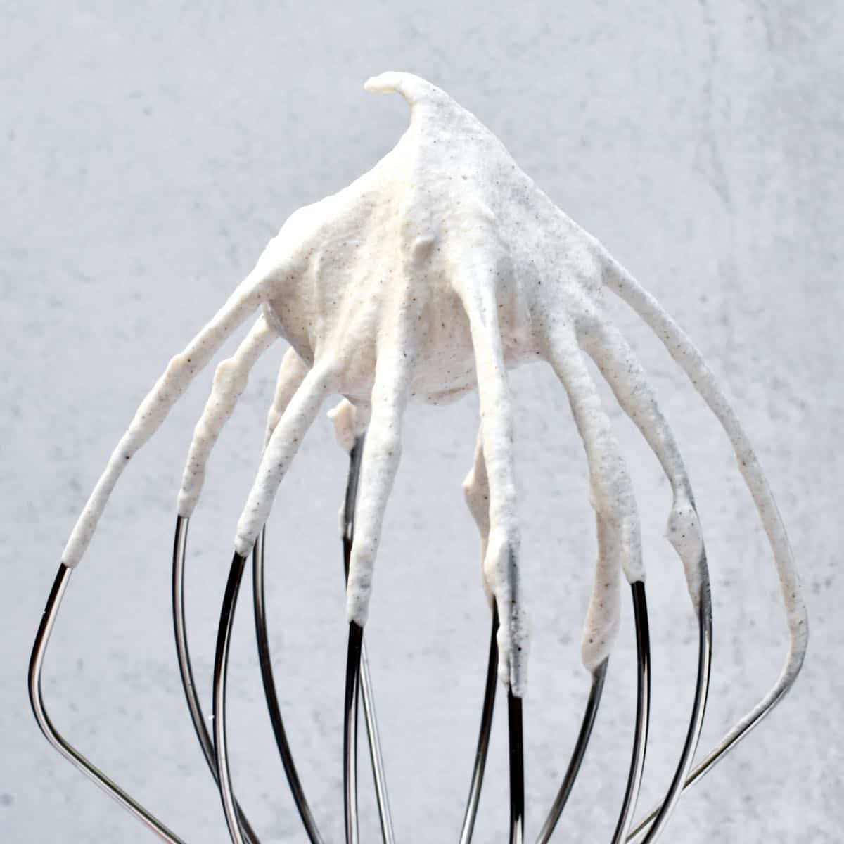 The whisk of a stand mixer with whipped cream showing a medium peak, curled at the tip.