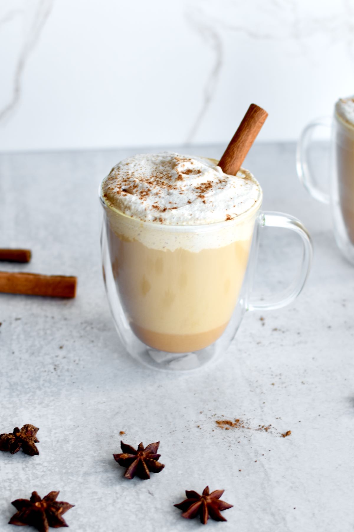 A glass coffee mug with a latte, topped with whipped cream and pumpkin spice. A cinnamon stick garnishes the latte, and star anise is sprinkled on the background.