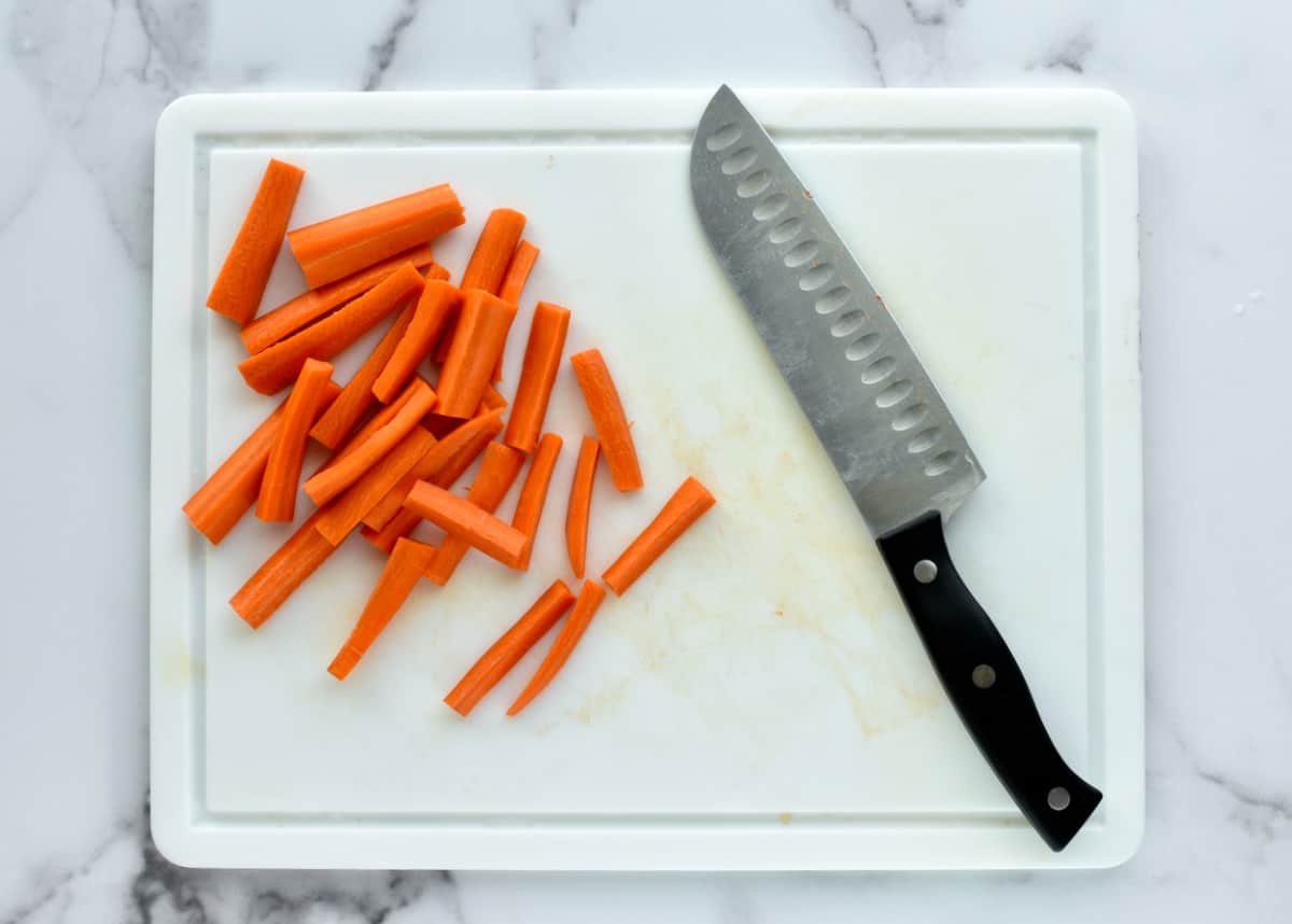 a white cutting board on a marble background with cut carrot sticks, next to a chef's knife.