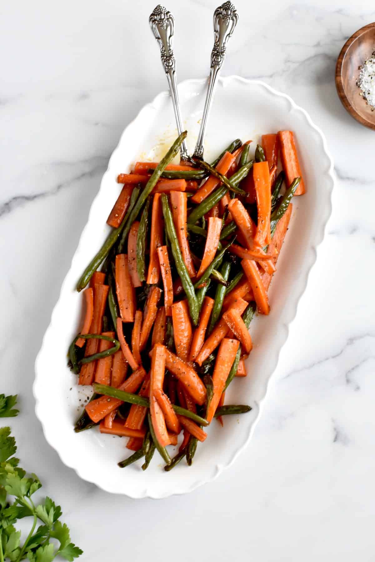 roasted carrots and green beans on a white scalloped dish on a marble background. A serving spoon and fork are resting in the dish, and a dish of salt and pepper and parsley is to the side.