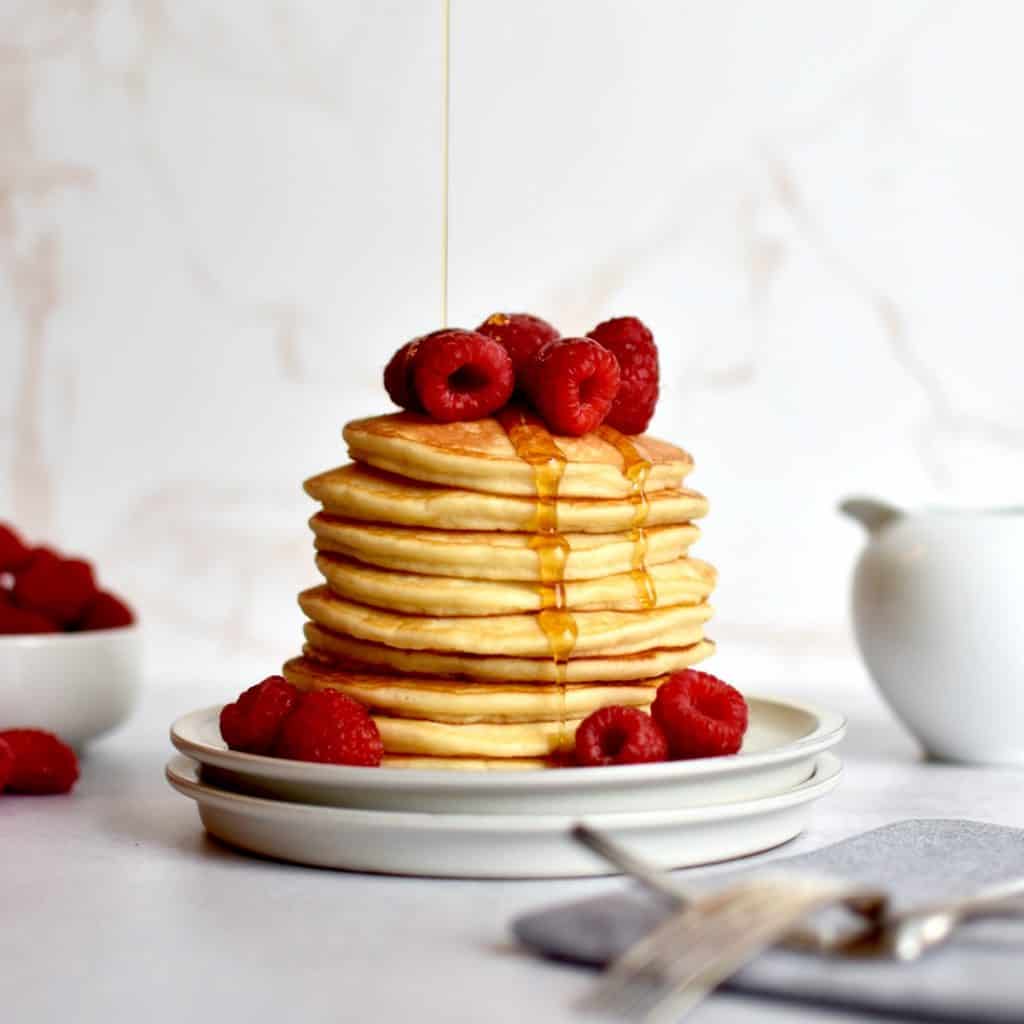 a stack of pancakes sitting on two plates, with a drizzle of syrup raining down on it. Raspberries are piled on top of the pancakes and two forks are resting in front of the plate on a napkin.