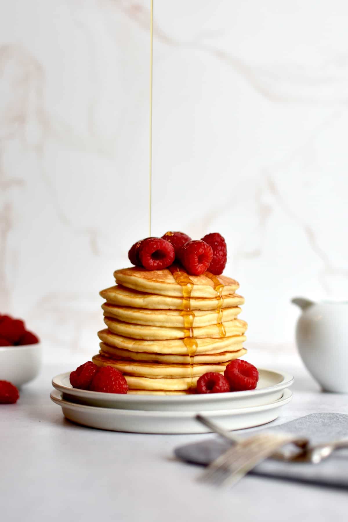 a stack of pancakes sitting on two plates, with a drizzle of syrup raining down on it. Raspberries are piled on top of the pancakes and two forks are resting in front of the plate on a napkin.