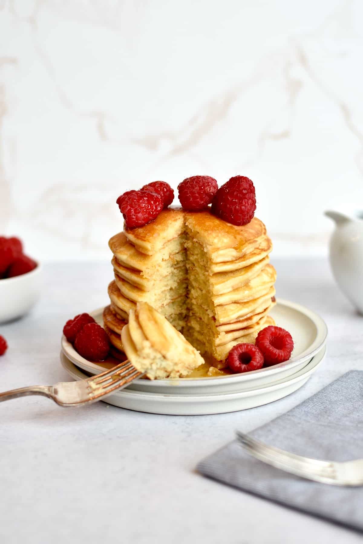 a stack of pancakes sitting on two plates, with a triangular cut out of the whole stack. Raspberries are piled on top of the pancakes and a fork is resting on the plate with a few pancake triangles speared on it.