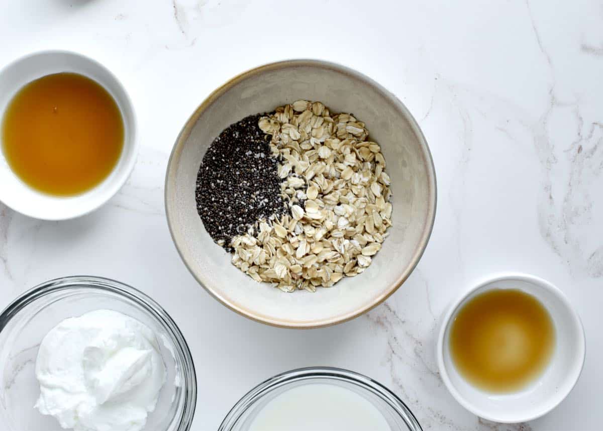 A grey bowl on a marble background with oats and chia seeds. Bowls containing yogurt, milk, syrup and vanilla are to the side.