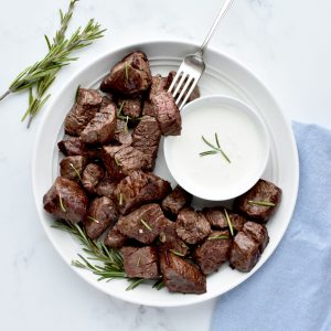 A white plate on a marble background. The plate holds steak bites, with a white bowl of horseradish sauce, and is garnished with rosemary. A fork spears into one steak bite.