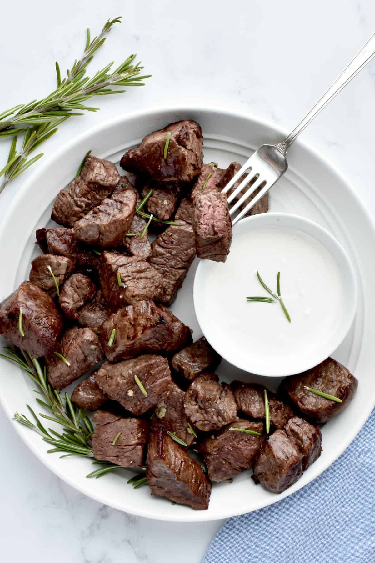 A white plate on a marble background. The plate holds steak bites, with a white bowl of horseradish sauce, and is garnished with rosemary. A fork spears into one steak bite.