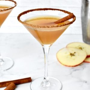 An apple cider martini garnished with a cinnamon stick on a marble background. A second martini and apple slices are in the background.