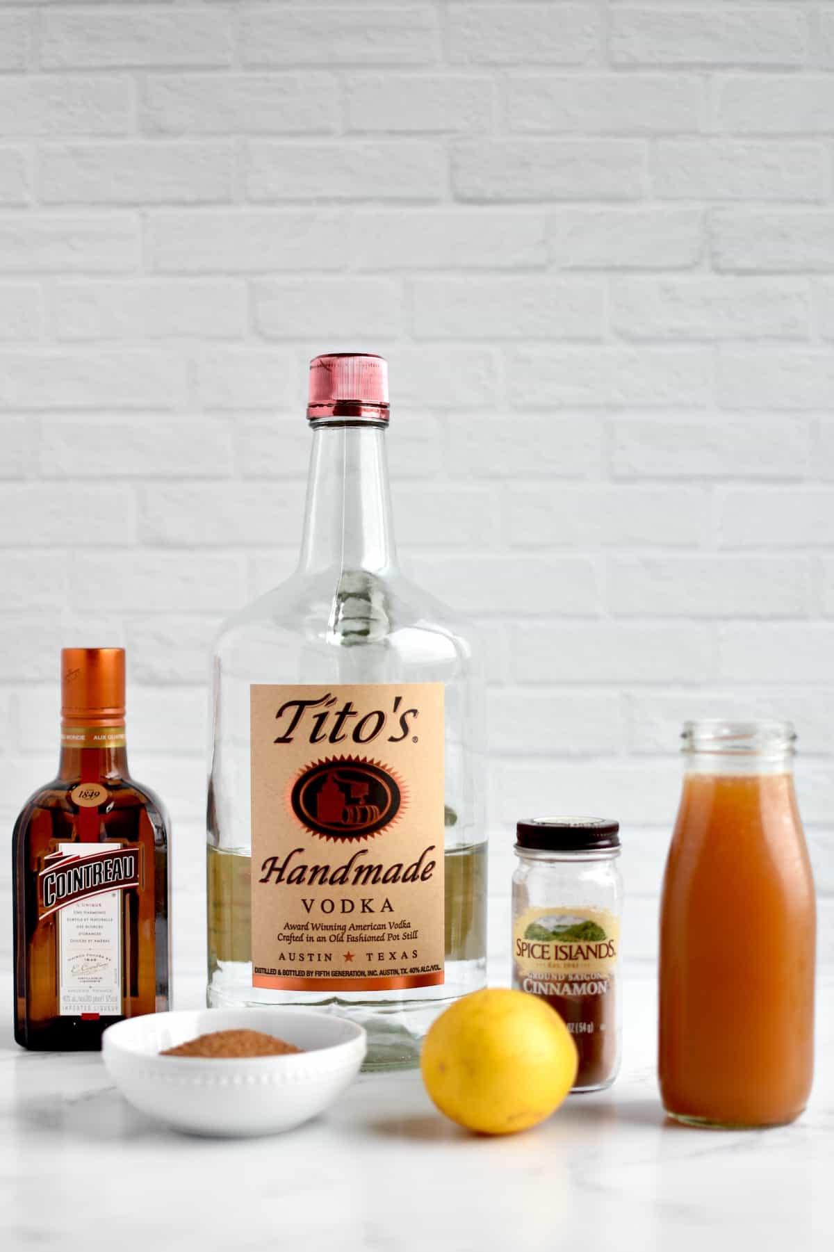 Ingredients for apple cider martini against a white brick background, including a bottle of Vodka, Cointreau, apple cider, a jar of cinnamon, bowl of brown sugar and a lemon.