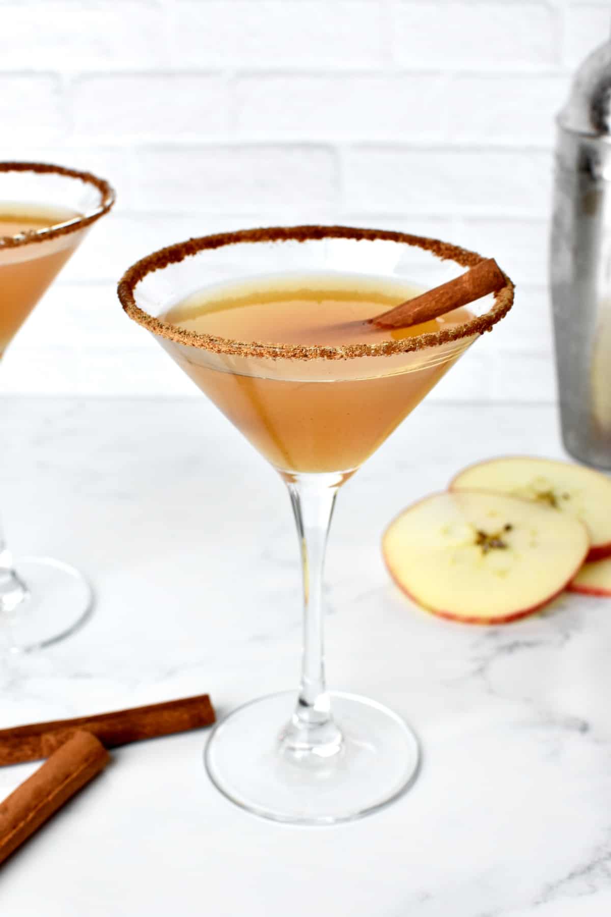 An apple cider martini garnished with a cinnamon stick on a marble background. A second martini and apple slices are in the background.