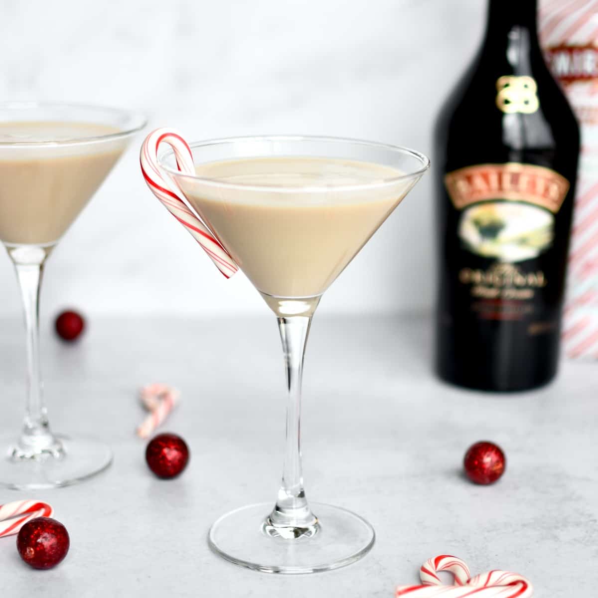 A martini glass on a grey counter, with a baileys espresso martini in it and a mini candy cane on the rim. A second martini, and bottles of baileys and peppermint vodka are in the background.