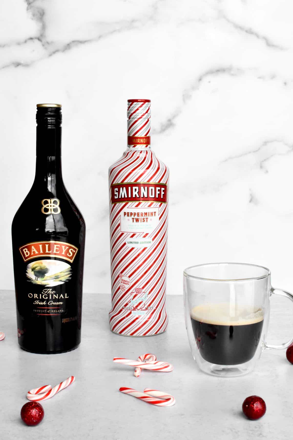 Ingredients for a bailey's peppermint espresso martini, including a bottle of baileys, a bottle of peppermint vodka, and a shot of espresso.