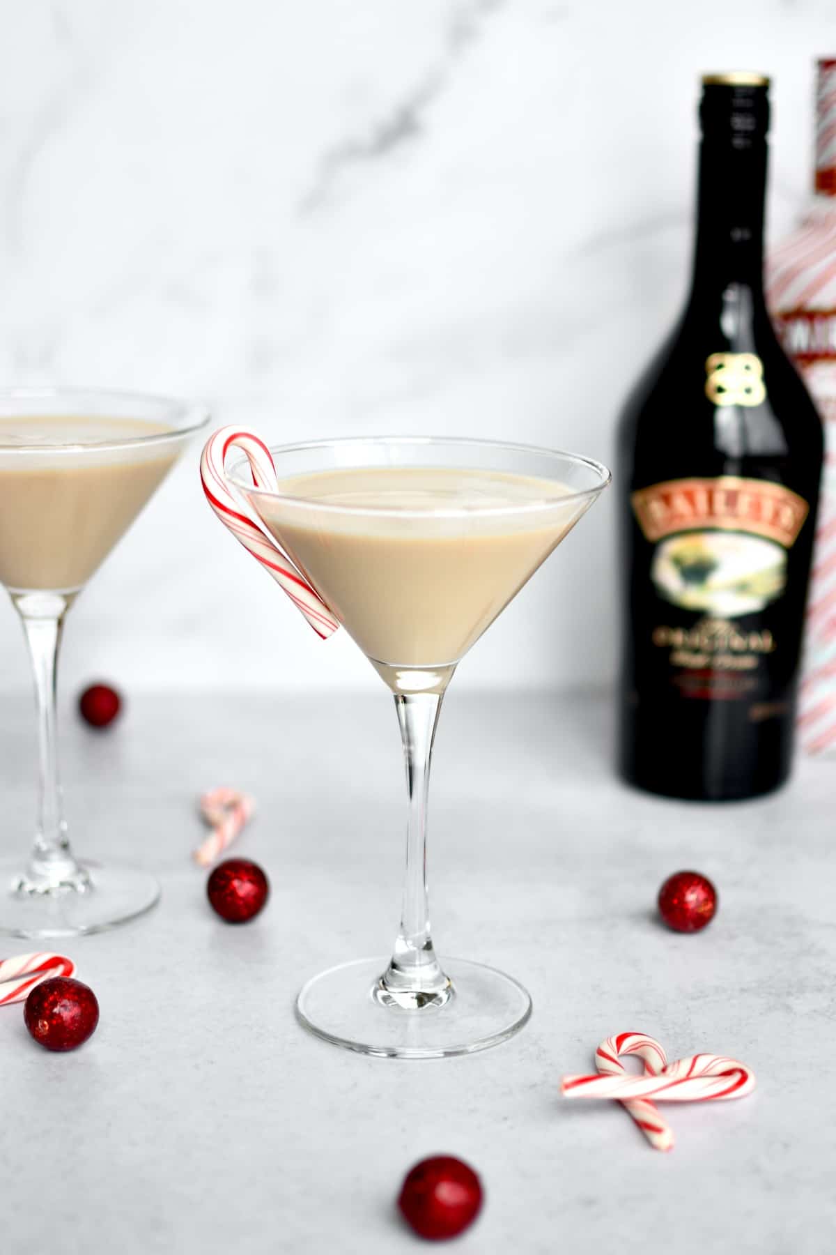 A martini glass on a grey counter, with a baileys espresso martini in it and a mini candy cane on the rim. A second martini, and bottles of baileys and peppermint vodka are in the background.