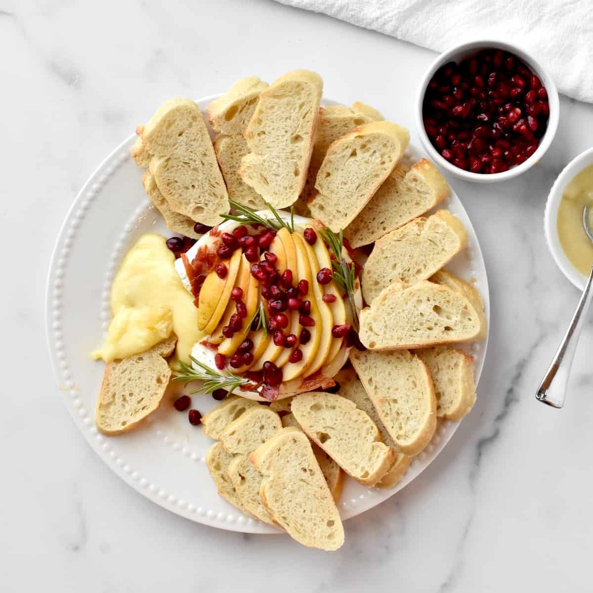 Top view of baked brie, topped with pears, prosciutto and pomegranate. The brie is surrounded by sliced bread and is oozing melted cheese.