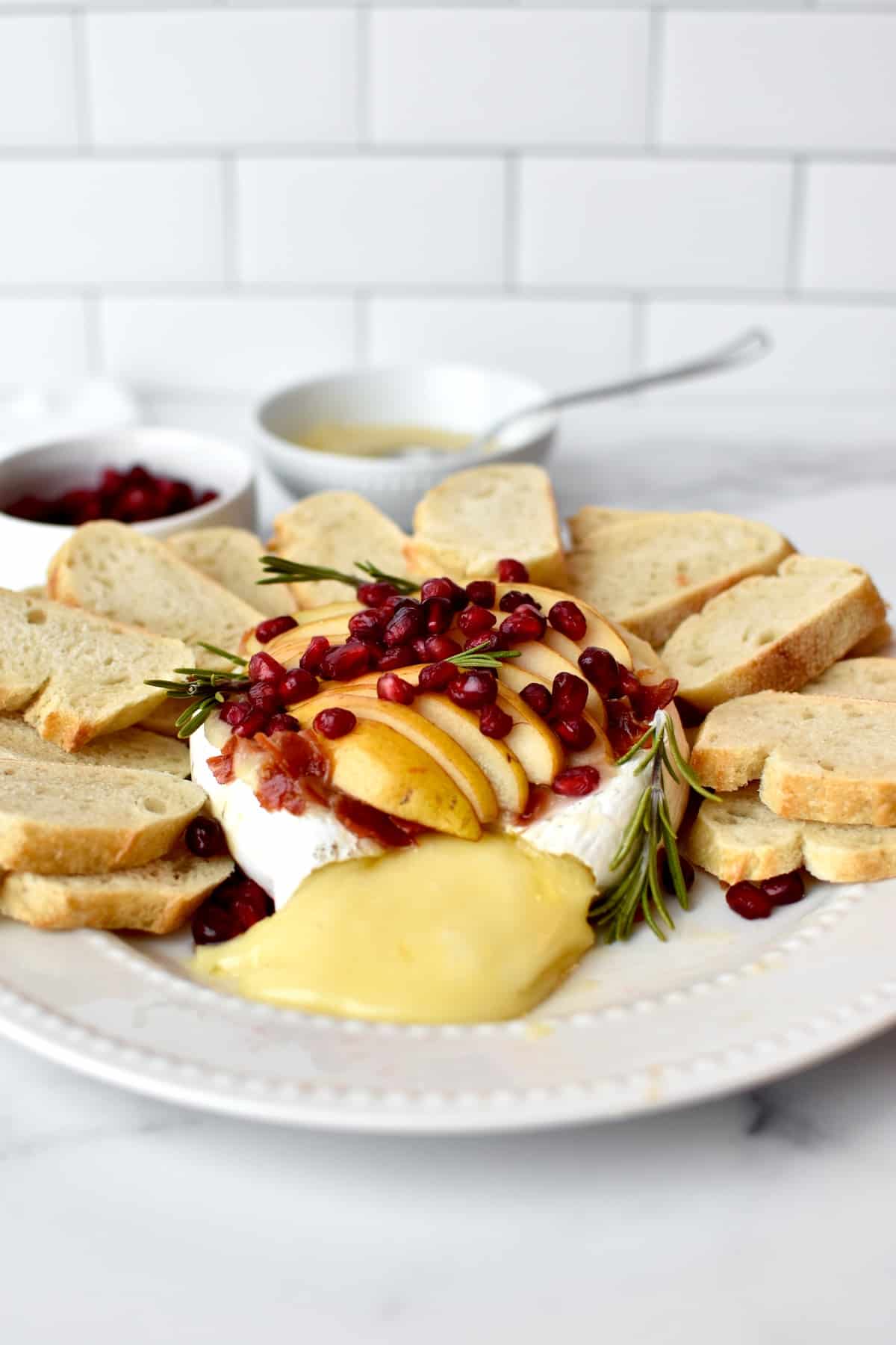 Side view of baked brie, topped with pears, prosciutto and pomegranate. The brie is surrounded by sliced bread and is oozing melted cheese.