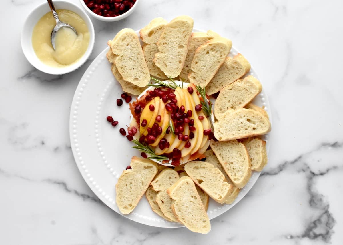 Top view of baked brie on a white plate, garnished with pomegranate and rosemary. The brie is surrounded by sliced bread.