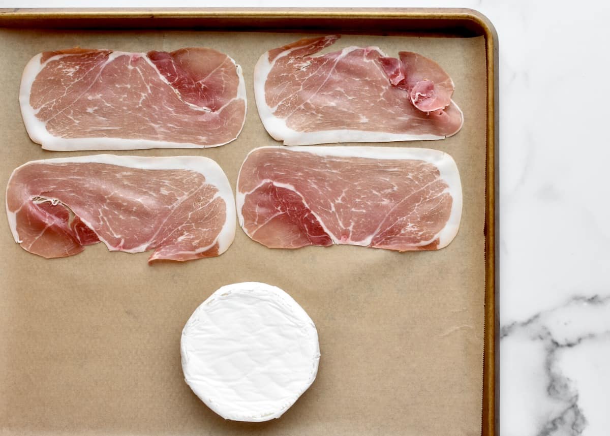 A sheet pan on a marble background. The sheet pan is lined with parchment paper and has a brie round and four slices of prosciutto on it.