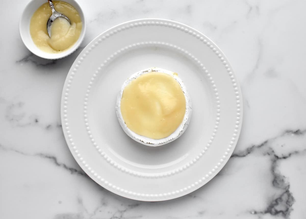 Baked brie on a white plate, sitting on a marble background. brie is topped with pear sauce.