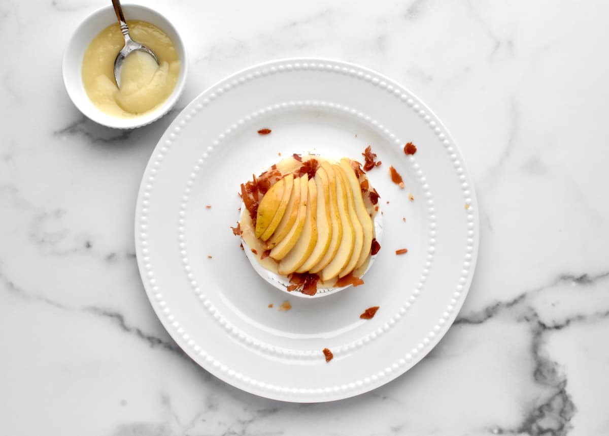 Baked brie on a white plate, sitting on a marble background. brie is topped with prosciutto and thinly sliced pears.