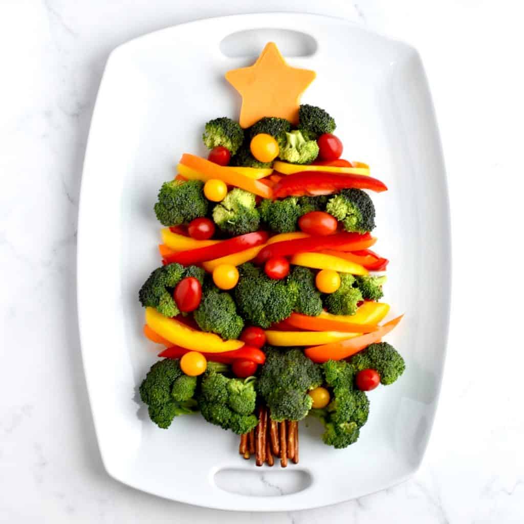 A holiday veggie tray on a white tray. Broccoli florets form a christmas tree shape, with bell pepper garland and cherry tomato ornaments.