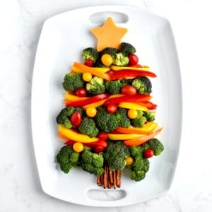 A holiday veggie tray on a white tray. Broccoli florets form a christmas tree shape, with bell pepper garland and cherry tomato ornaments.