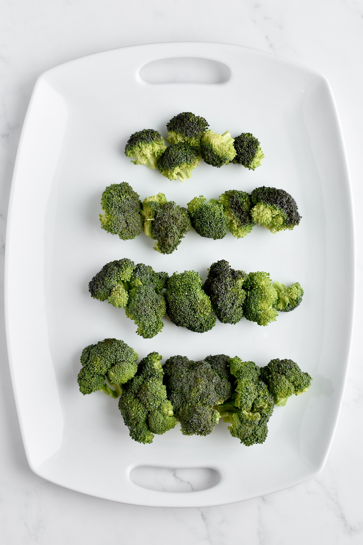 A white platter on a marble background showing four tiers of broccoli arranged to form a christmas tree.