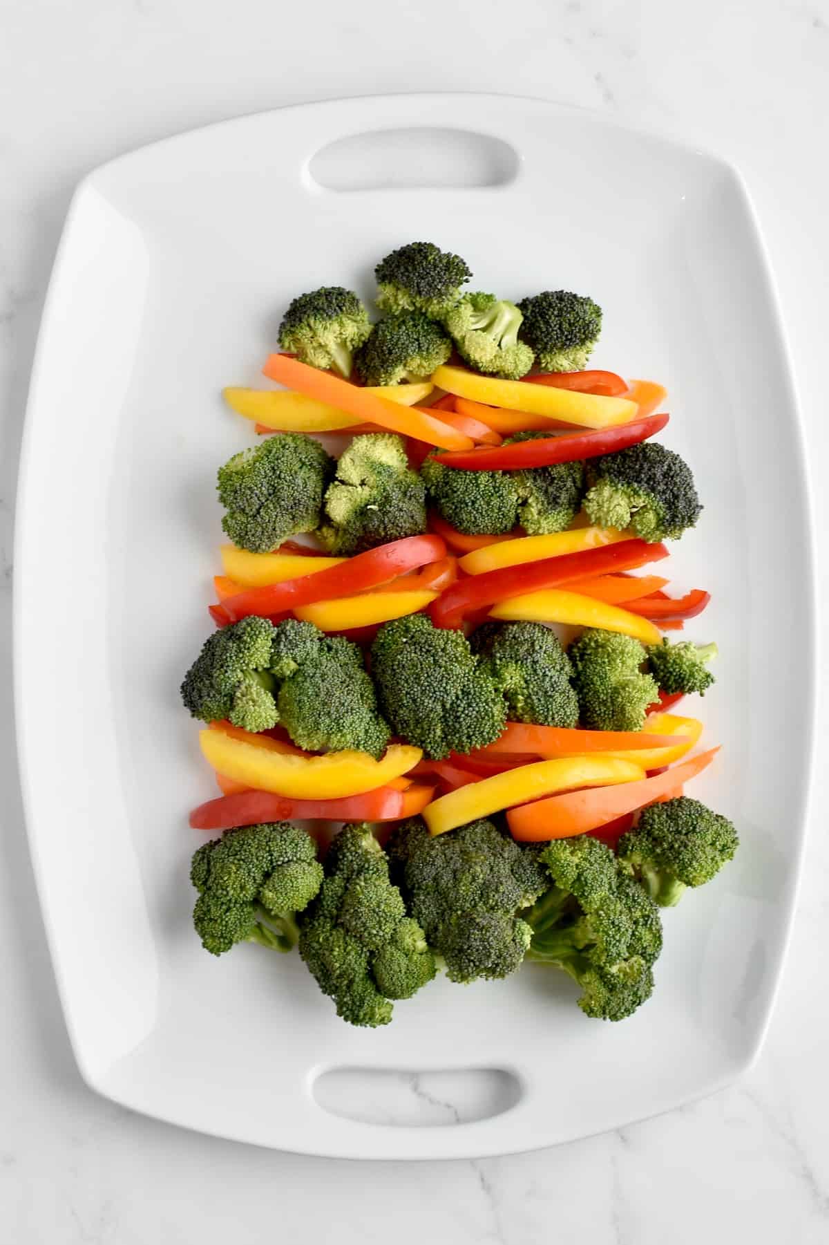 A white platter on a marble background showing four tiers of broccoli arranged to form a christmas tree, with bell pepper strips between each tier forming colorful garland.