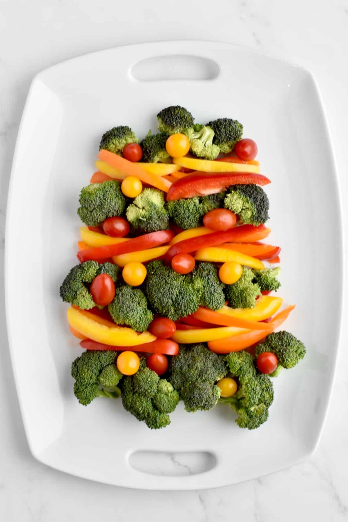 A white platter on a marble background showing four tiers of broccoli arranged to form a christmas tree, with bell pepper strips between each tier forming colorful garland and cherry tomatoes arranged as ornaments.