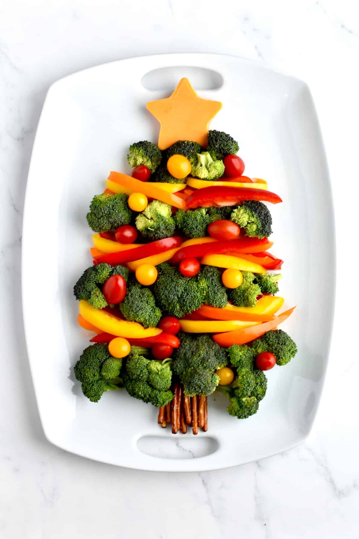 A holiday veggie tray on a white tray. Broccoli florets form a christmas tree shape, with bell pepper garland and cherry tomato ornaments.