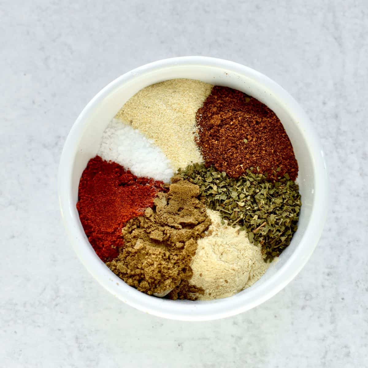 A white bowl on a grey background. The bowl contains 7 different spices, not yet mixed, so you can see all the distinct colors.