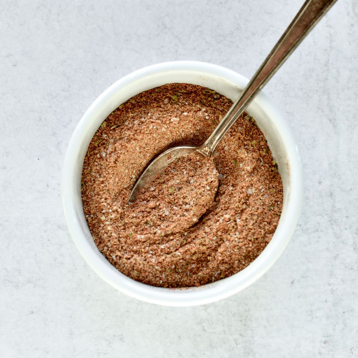 A white bowl on a grey background. The bowl contains mixed chicken taco seasoning being stirred with a spoon.