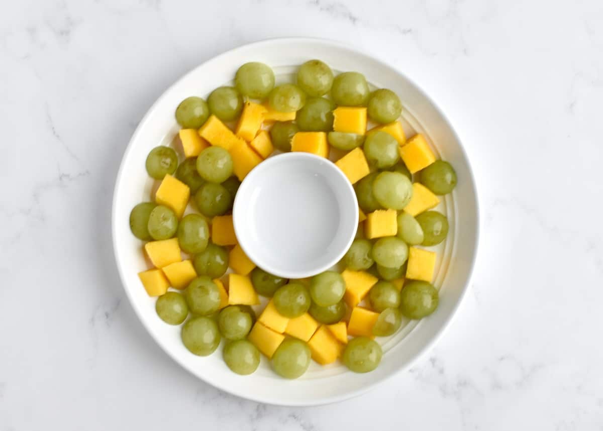 The first layer of a fruit salad, with yellow mangos and green grapes arranged in a wreath shape.