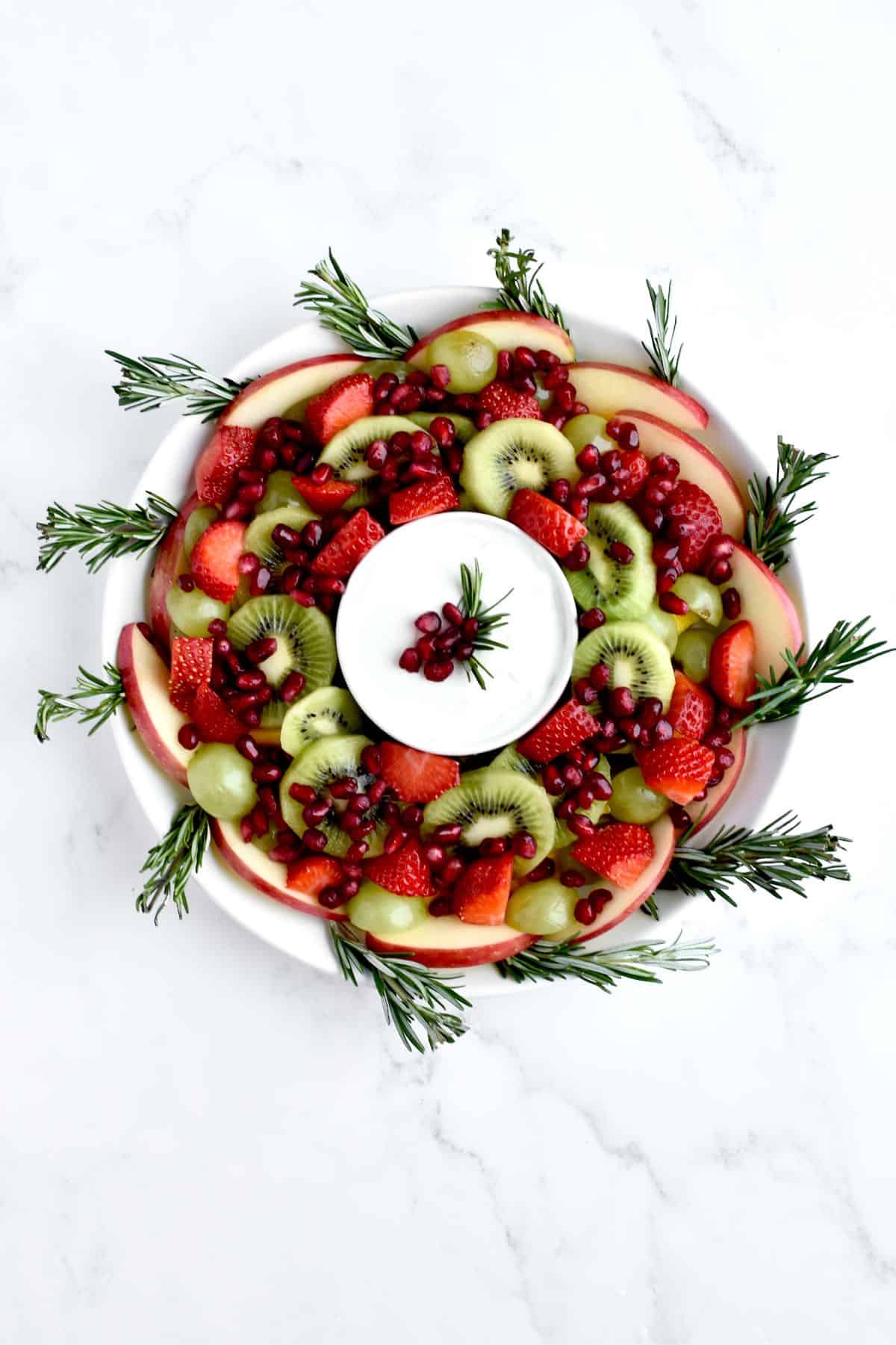 A fruit salad arranged in a wreath shape on a white plate, with a bowl of yogurt sauce in the middle.