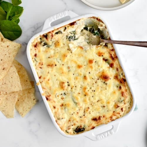 A white casserole dish with crab spinach artichoke on a marble background, with tortilla chips and spinach at the side.